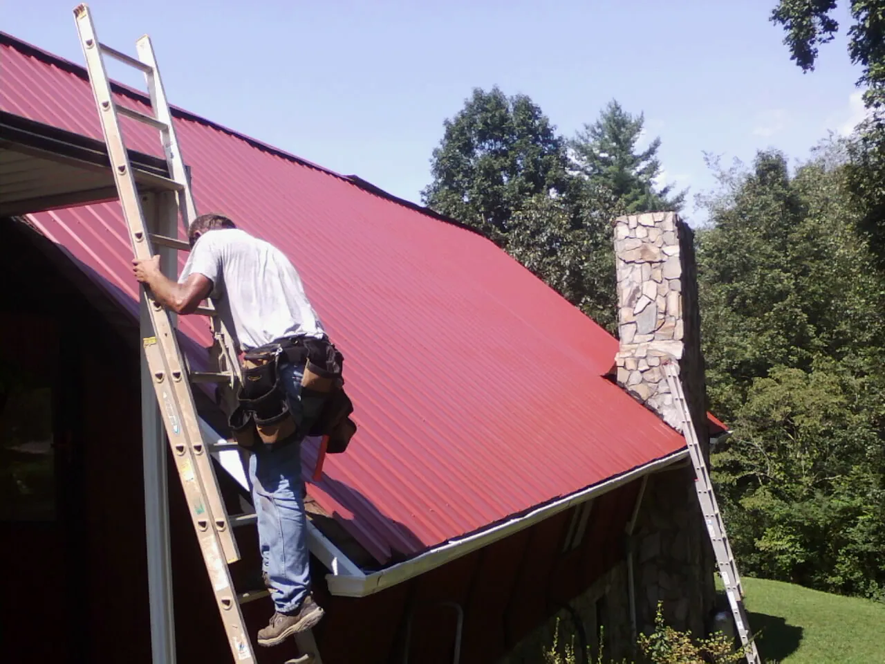 Roofer on a ladder beside a red metal roof and stone chimney — Soffit & Fascia Repair by Roofing Brothers in South Sterling
